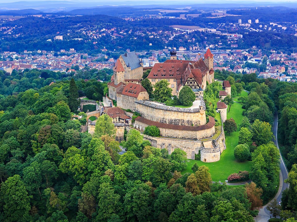 Majestätische historische Burg auf einem grünen Hügel, umgeben von dichtem Wald und einer Stadt im Hintergrund.