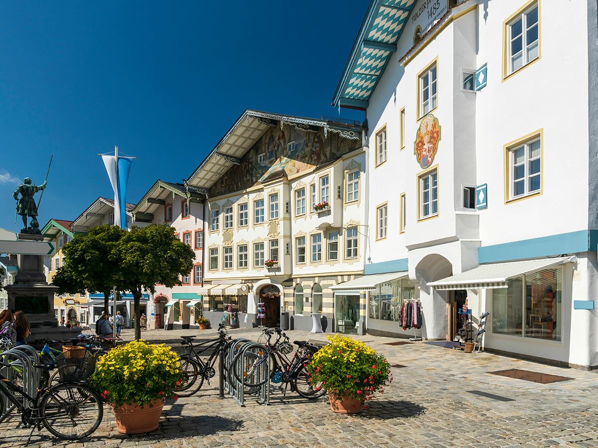 Sonniger Marktplatz in Bad Tölz, Bayern, mit traditionellen, bemalten Gebäuden, Statue, Fahrrädern und Blumenarrangements.