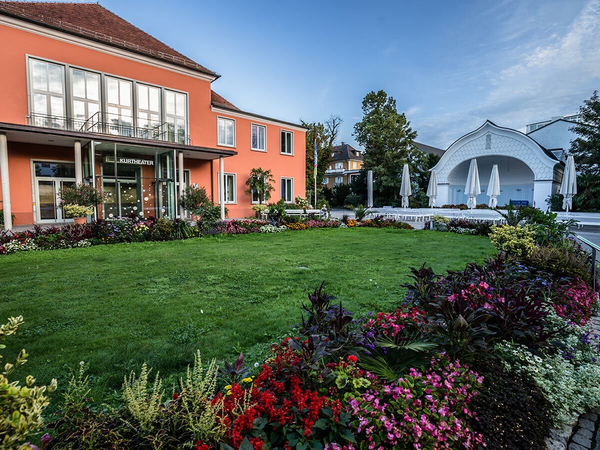 Das Kurtheater mit farbenfrohen Blumen, grünem Rasen und einer Freilichtbühne im Hintergrund an einem hellen Tag.
