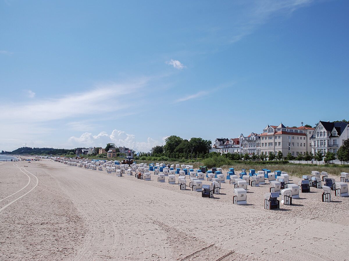 Zahlreiche Strandkörbe stehen auf einem sonnigen Sandstrand vor historischen Gebäuden eines deutschen Seebades.