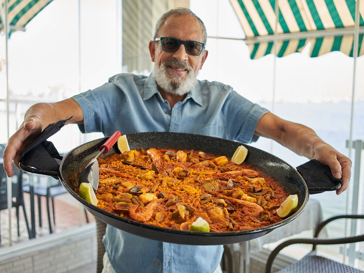 Ein lächelnder, bärtiger Mann mit Brille präsentiert stolz eine große, traditionelle Meeresfrüchte-Paella im Freien.