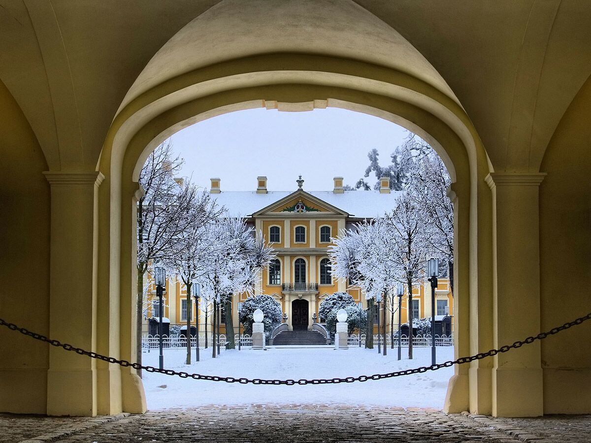 Historisches gelbes Schloss im Winter, umrahmt von einem Torbogen, mit schneebedeckten Bäumen und Boden.