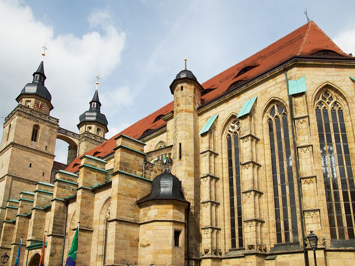 Historische Steinkirche mit markanten Zwillingstürmen, Bogenbrücke und gotischen Fenstern unter bewölktem Himmel.