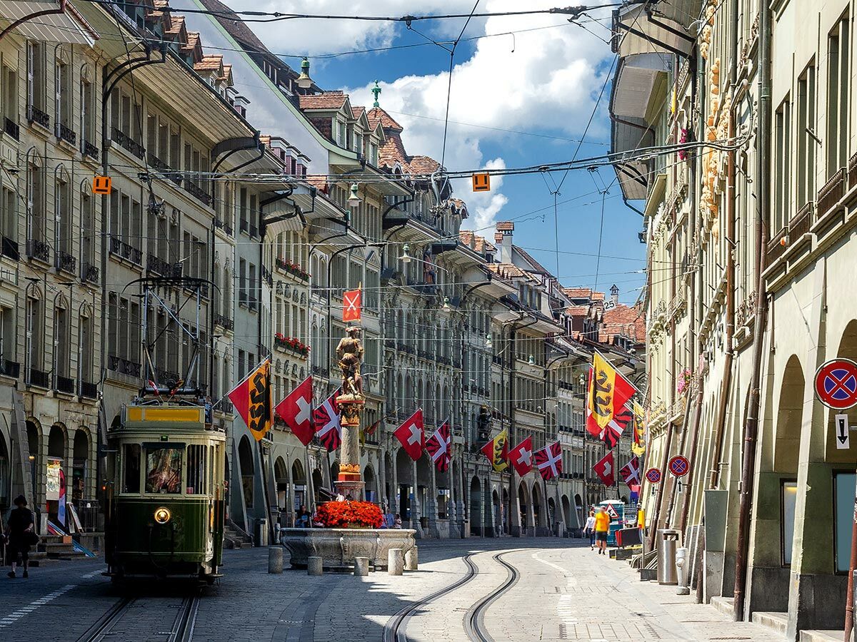 Malerische Bern Altstadt mit historischer grüner Straßenbahn, Zähringerbrunnen und Schweizer Flaggen unter blauem Himmel.