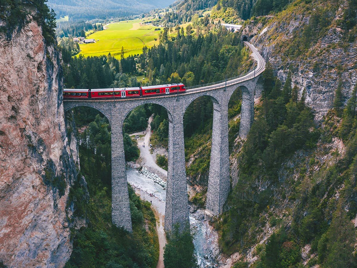 Roter Bernina Express Zug fährt über das historische Landwasser Viadukt in einer tiefen Schlucht mit Fluss, Wald und Bergen in der Schweiz.