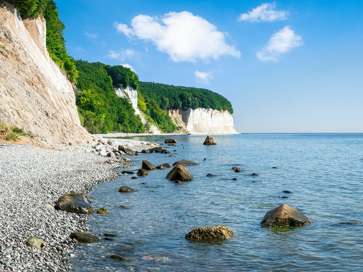 Blick auf die Kreidefelsen von Rügen an der Ostsee mit grünem Wald, Felsstrand und klarem blauen Meer unter sonnigem Himmel.