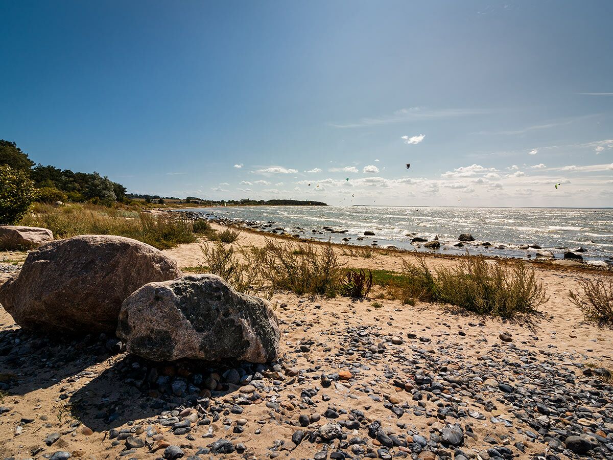 Sonnenüberfluteter Strand mit großen Felsen, Kies und Sand am Ufer, dahinter das glitzernde Meer mit Kitesurfern unter blauem Himmel.