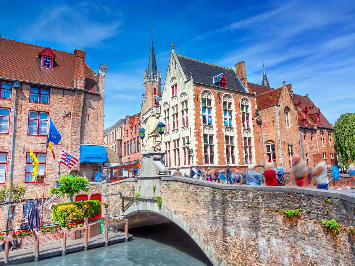 Historische Altstadt von Brügge mit Kanal, Steinbrücke, traditionellen Gebäuden und Menschen unter blauem Himmel.