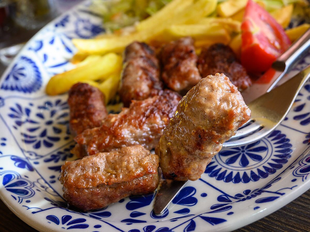 Ein Teller mit gegrillten Cevapcici, goldbraunen Pommes frites und einer Tomatenscheibe, ein Stück am Gabel.