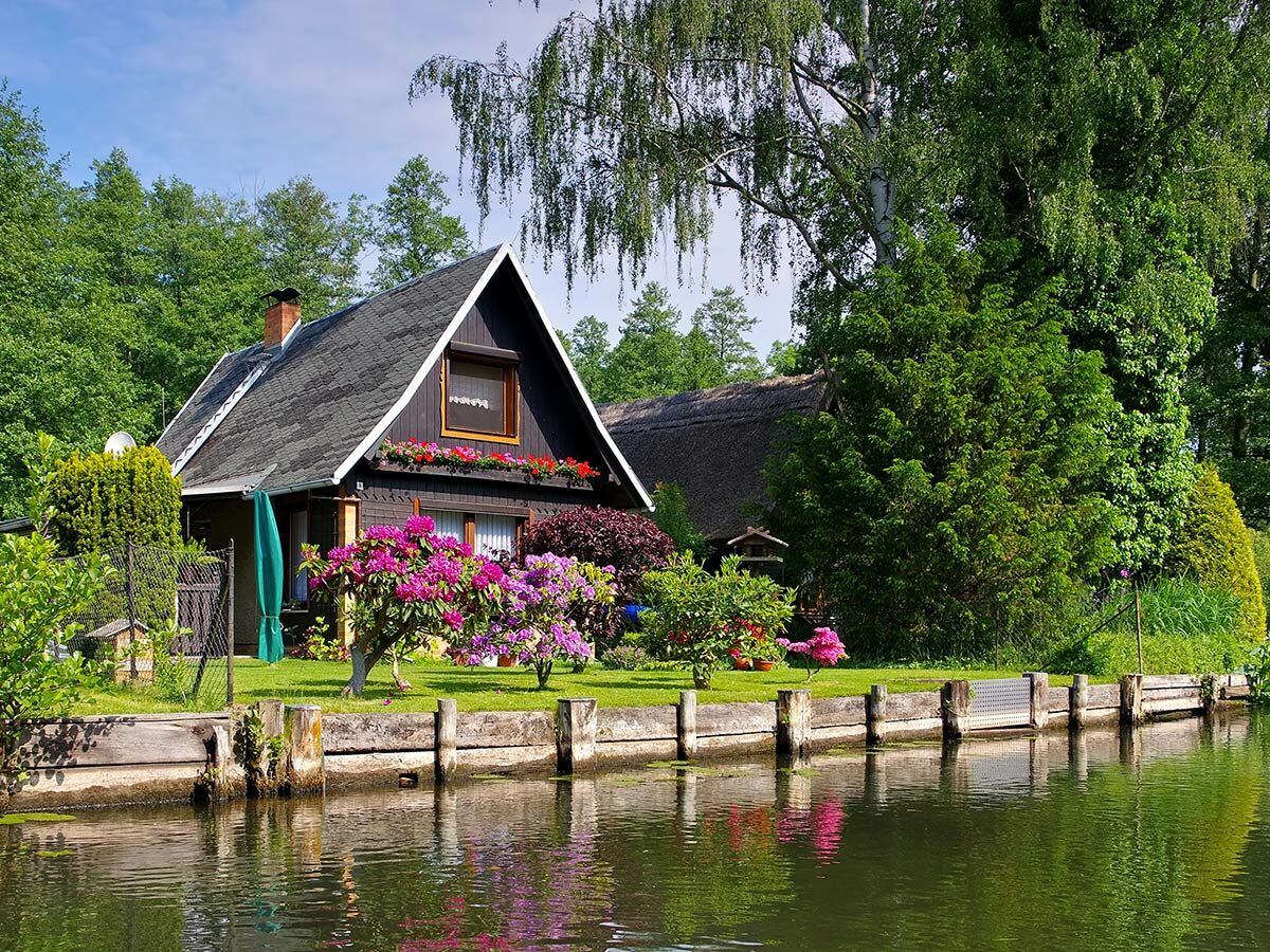 Malerisches Haus am Kanal mit bunten Blumen, grünem Rasen und Wasserspiegelungen an einem sonnigen Tag.