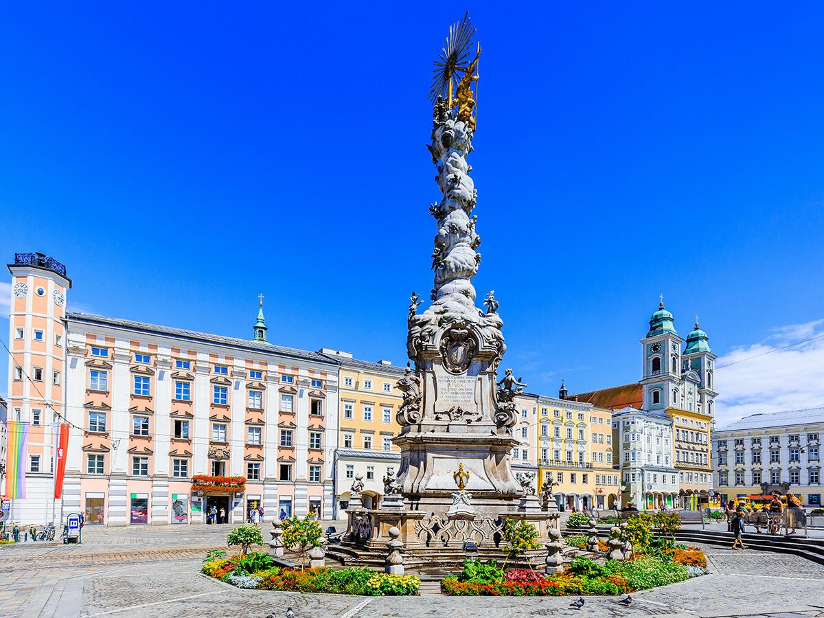 Barocke Pestsäule auf dem Hauptplatz in Linz, Österreich, mit historischen Gebäuden und blauem Himmel im Hintergrund.