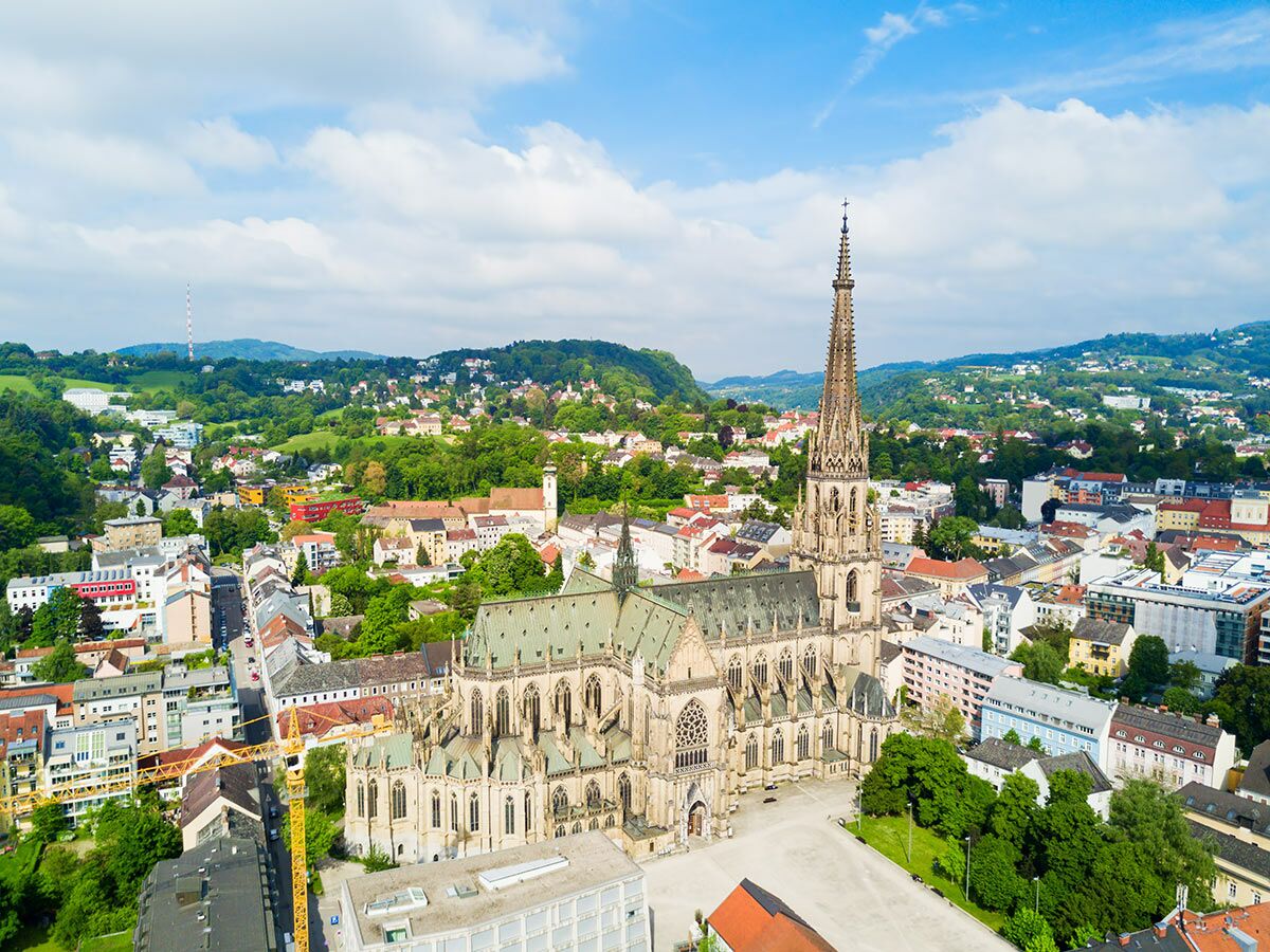 Luftbild der gotischen Minoritenkirche in Wels, Österreich, umgeben von Stadtgebäuden und grünen Hügeln bei Tag mit blauem Himmel. Baustelle im Vordergrund.