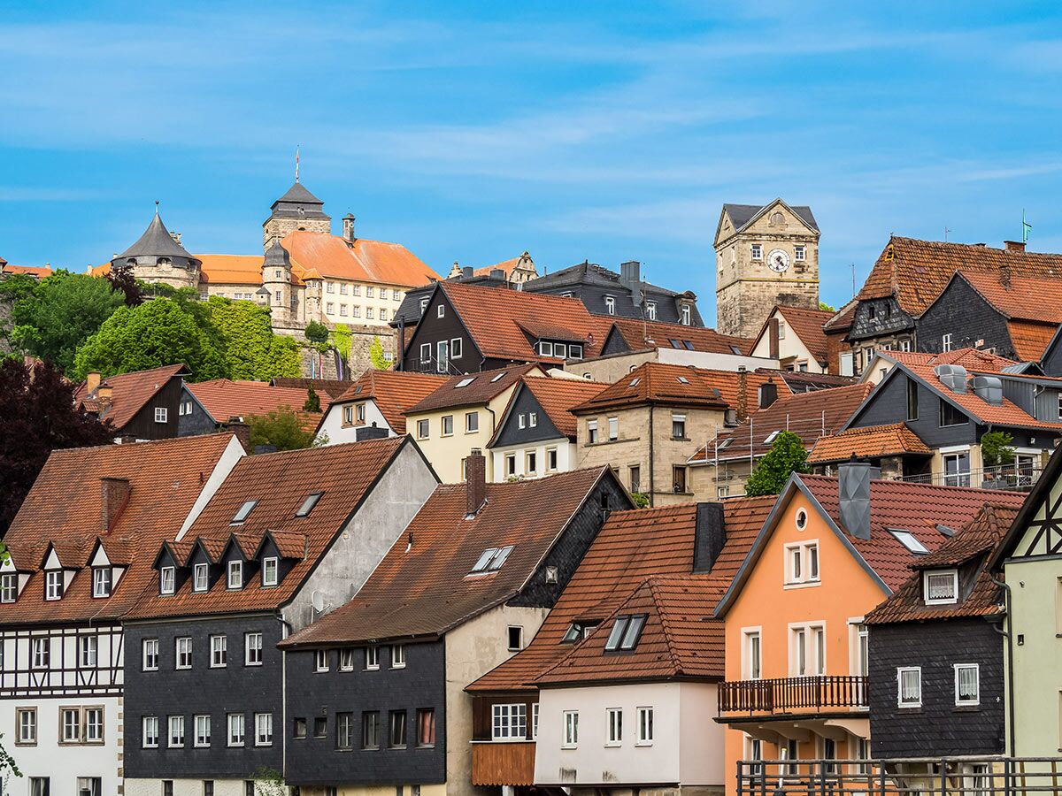 Malerische historische Stadtansicht mit einer Burg auf dem Hügel, einem Uhrturm und vielen Häusern mit roten Dächern unter blauem Himmel.