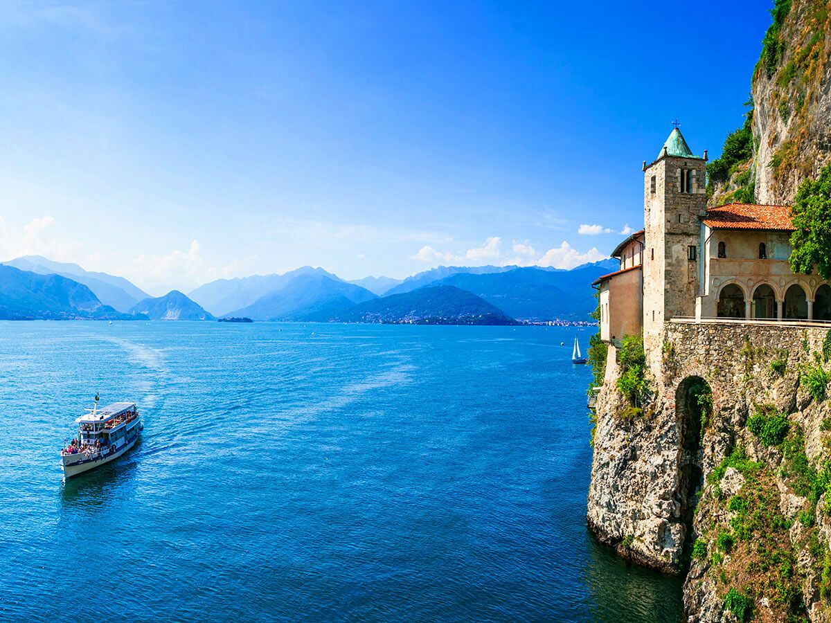 Historisches Kloster Santa Caterina del Sasso am Lago Maggiore mit Passagierschiff, Segelboot und Alpenpanorama.