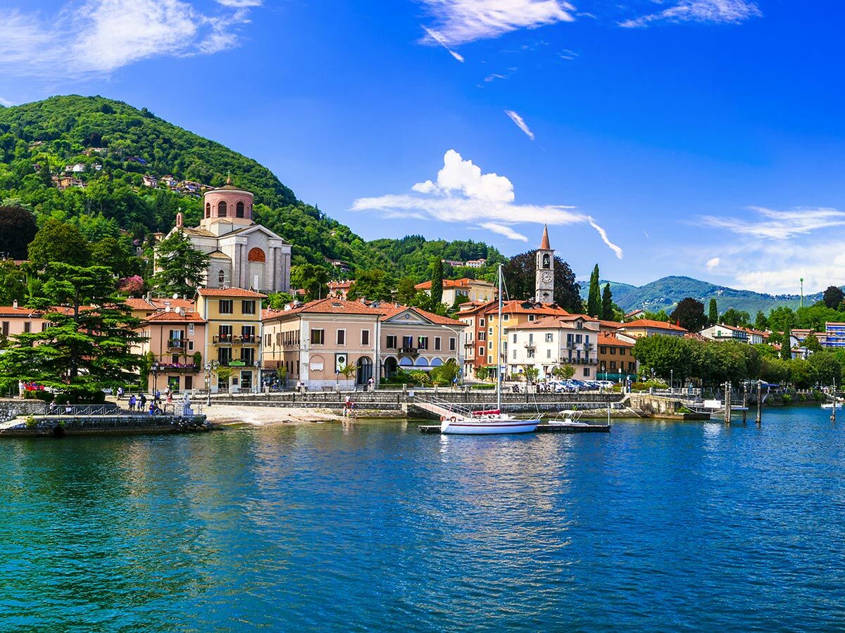 Malerischer italienischer Seeuferort mit bunten Häusern, Kirchen, grünen Bergen und blauer See mit Segelbooten unter blauem Himmel.
