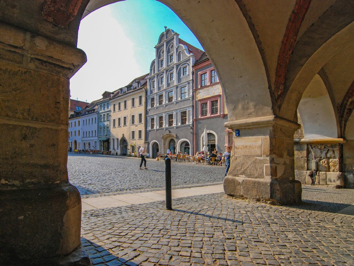 Blick durch Steinbögen auf Görlitzer Untermarkt mit Kopfsteinpflaster, farbigen Giebelhäusern, Straßencafés und Menschen.