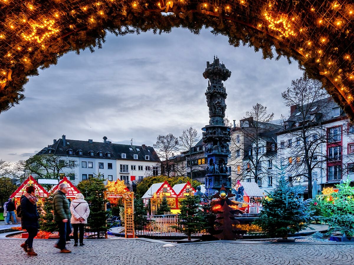 Festlicher Weihnachtsmarkt in der Dämmerung mit Lichtern, Menschen, Ständen und einem prominenten historischen Denkmal auf einem Stadtplatz.