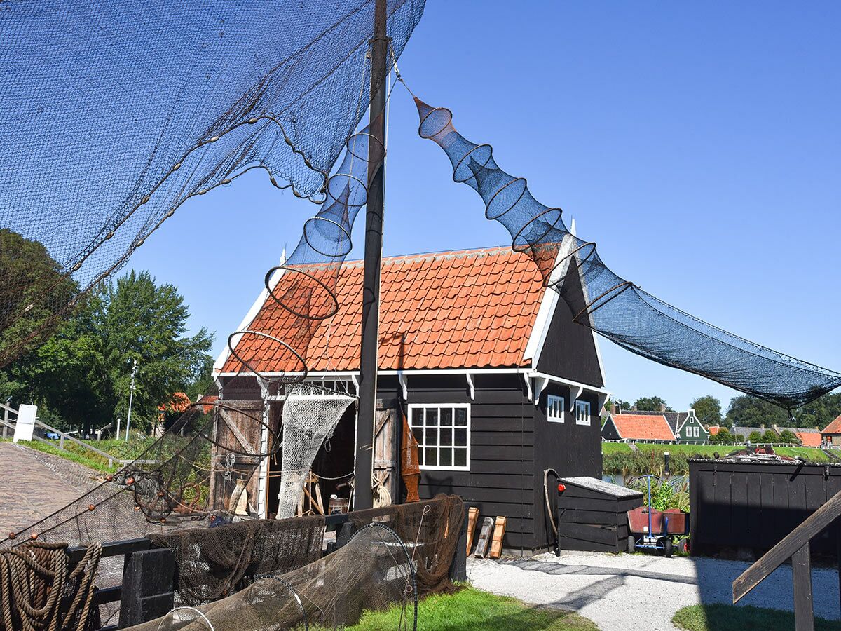 Traditionelles schwarzes Fischerhaus mit rotem Dach, davor großes Fischernetz an einem Pfahl zum Trocknen ausgebreitet unter blauem Himmel.
