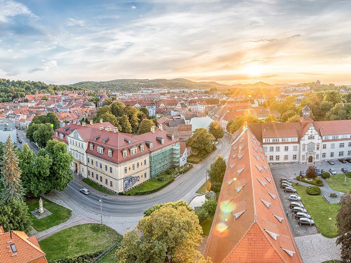Luftaufnahme einer historischen europäischen Stadt bei Sonnenuntergang mit roten Dächern, alten Gebäuden, grünen Bäumen und hügeliger Landschaft.