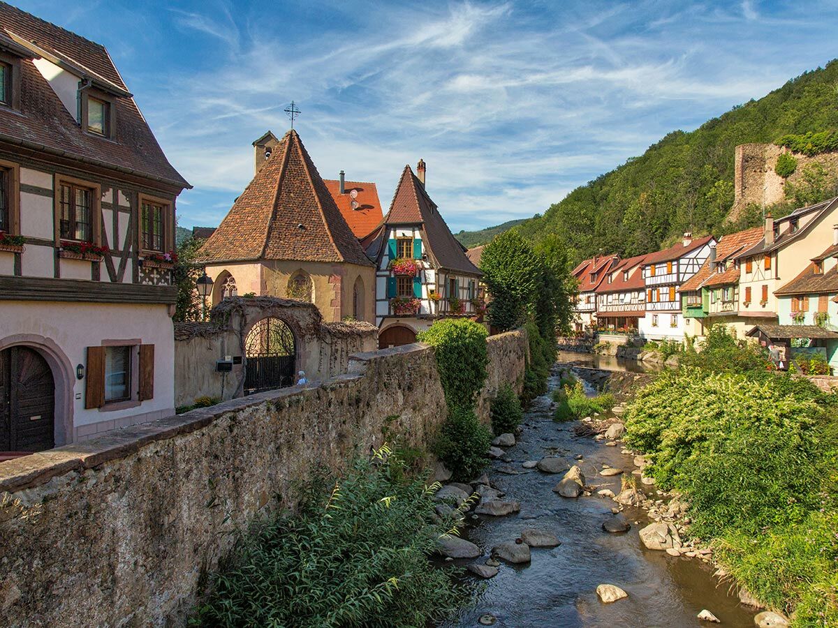 Malerisches Dorf am Fluss in Elsass mit Fachwerkhäusern, alter Kirche, Steinmauer, grünen Hügeln und Burgruine.