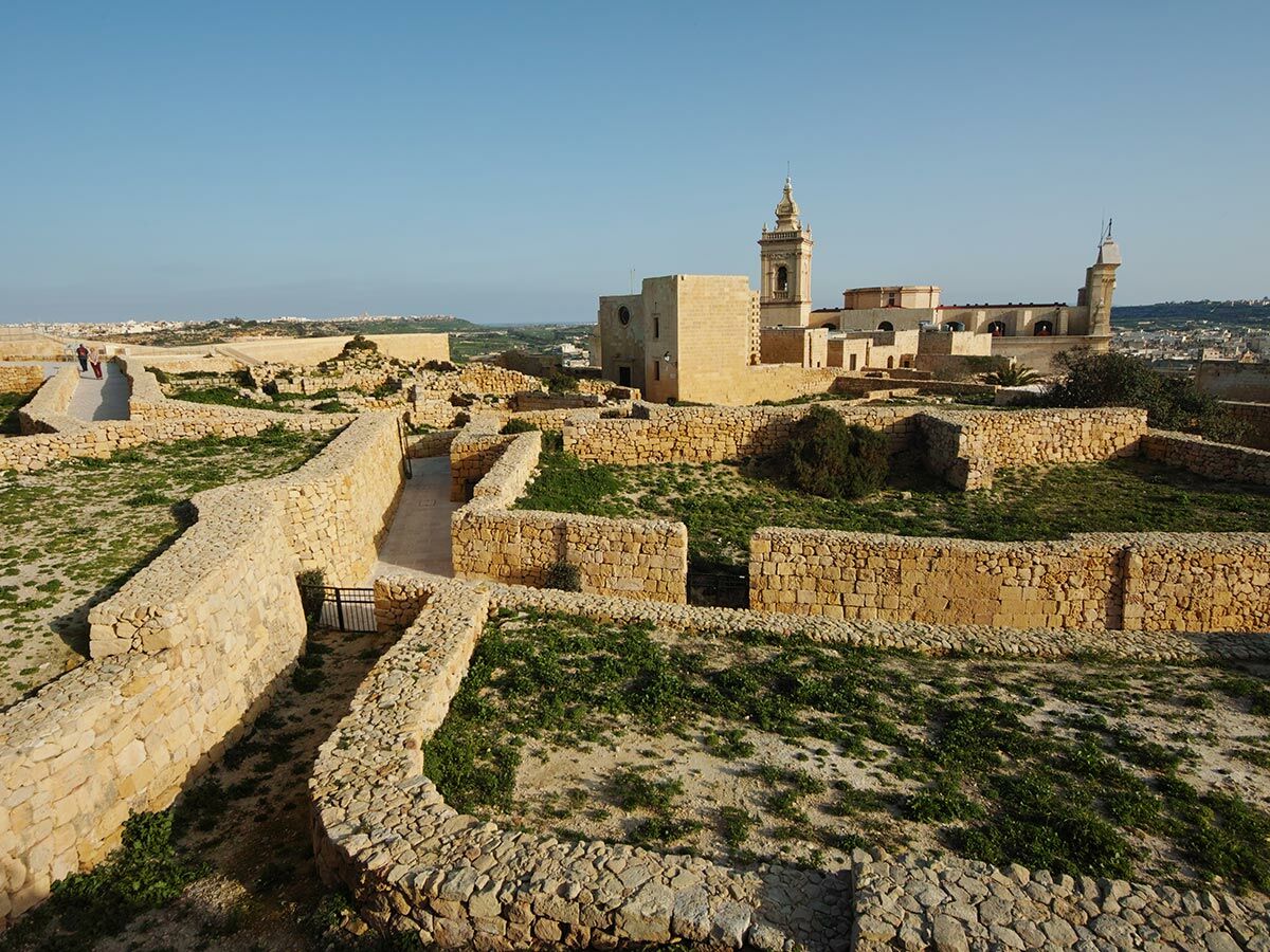 Blick auf die historische Zitadelle auf Gozo mit alten Steinmauern, grünen Flächen und der Kathedrale mit Glockenturm unter blauem Himmel.