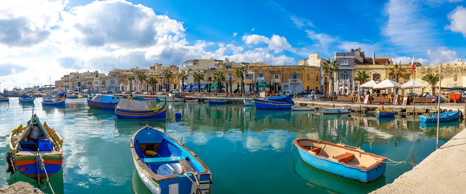 Bunte Luzzu-Fischerboote schwimmen im türkisfarbenen Hafen von Marsaxlokk, Malta, mit Uferpromenade und Gebäuden.