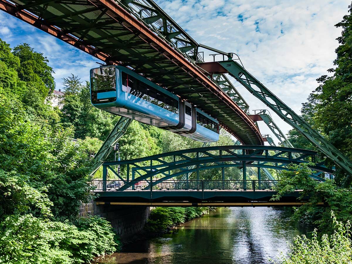 Blaue Wuppertaler Schwebebahn fährt über den Fluss Wupper, umgeben von grünen Bäumen und einer Straßenbrücke.