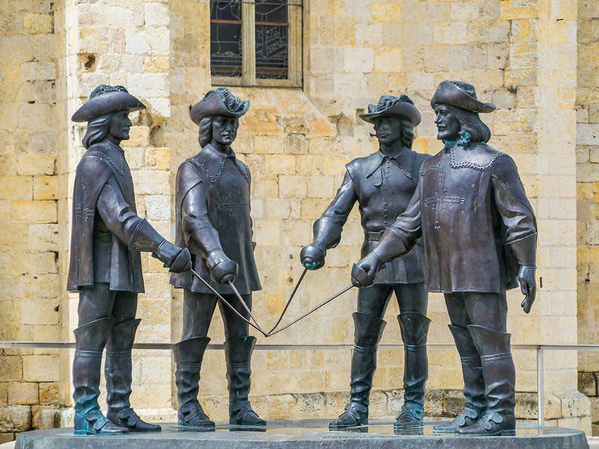 Vier Bronzestatuen historischer Männer mit Hüten und Schwertern stehen vor einer alten Steinmauer mit Fenster.