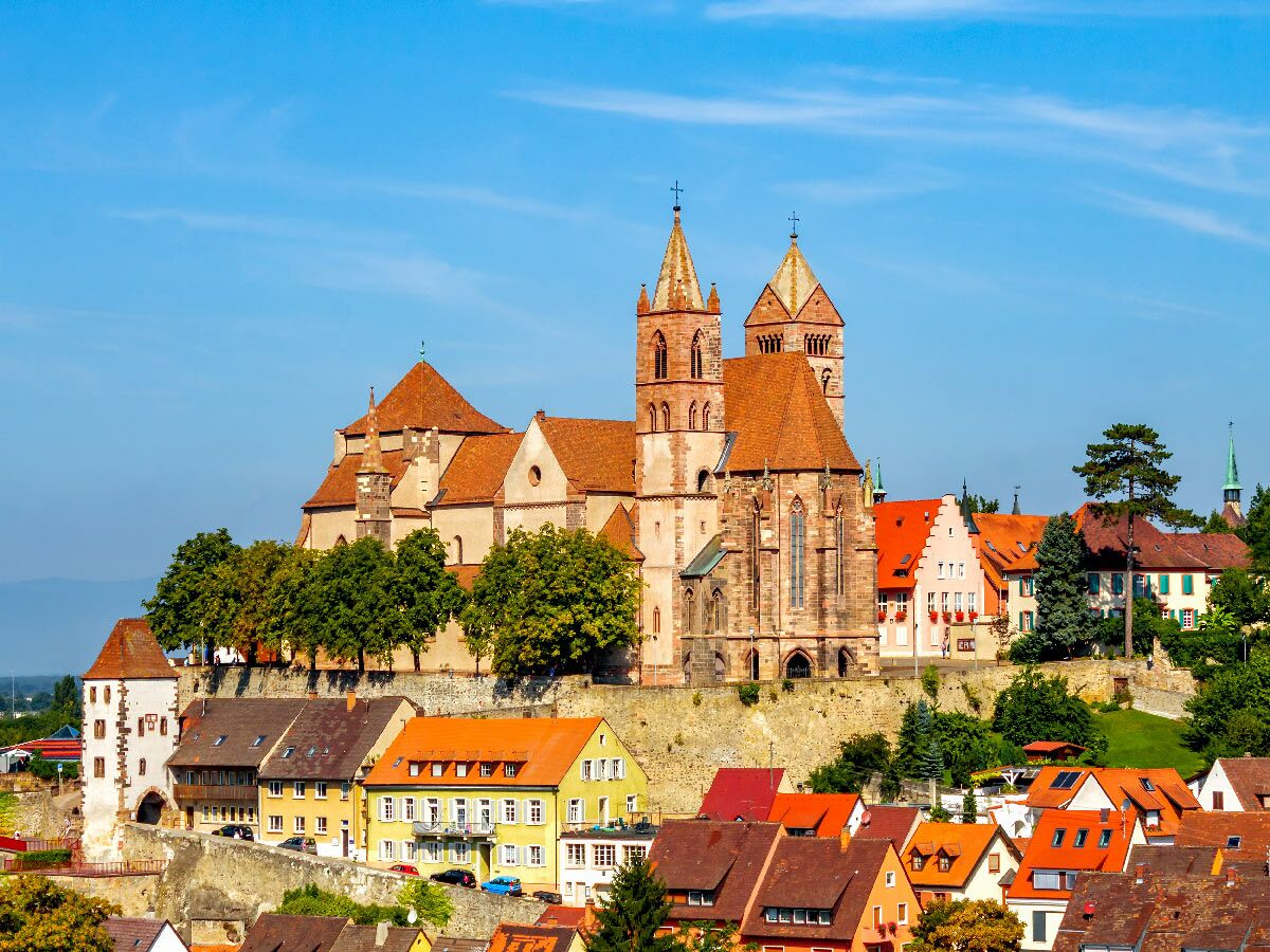 Blick auf eine malerische europäische Altstadt mit großem romanischen Münster auf einem Hügel, umgeben von Häusern mit roten Dächern unter blauem Himmel.