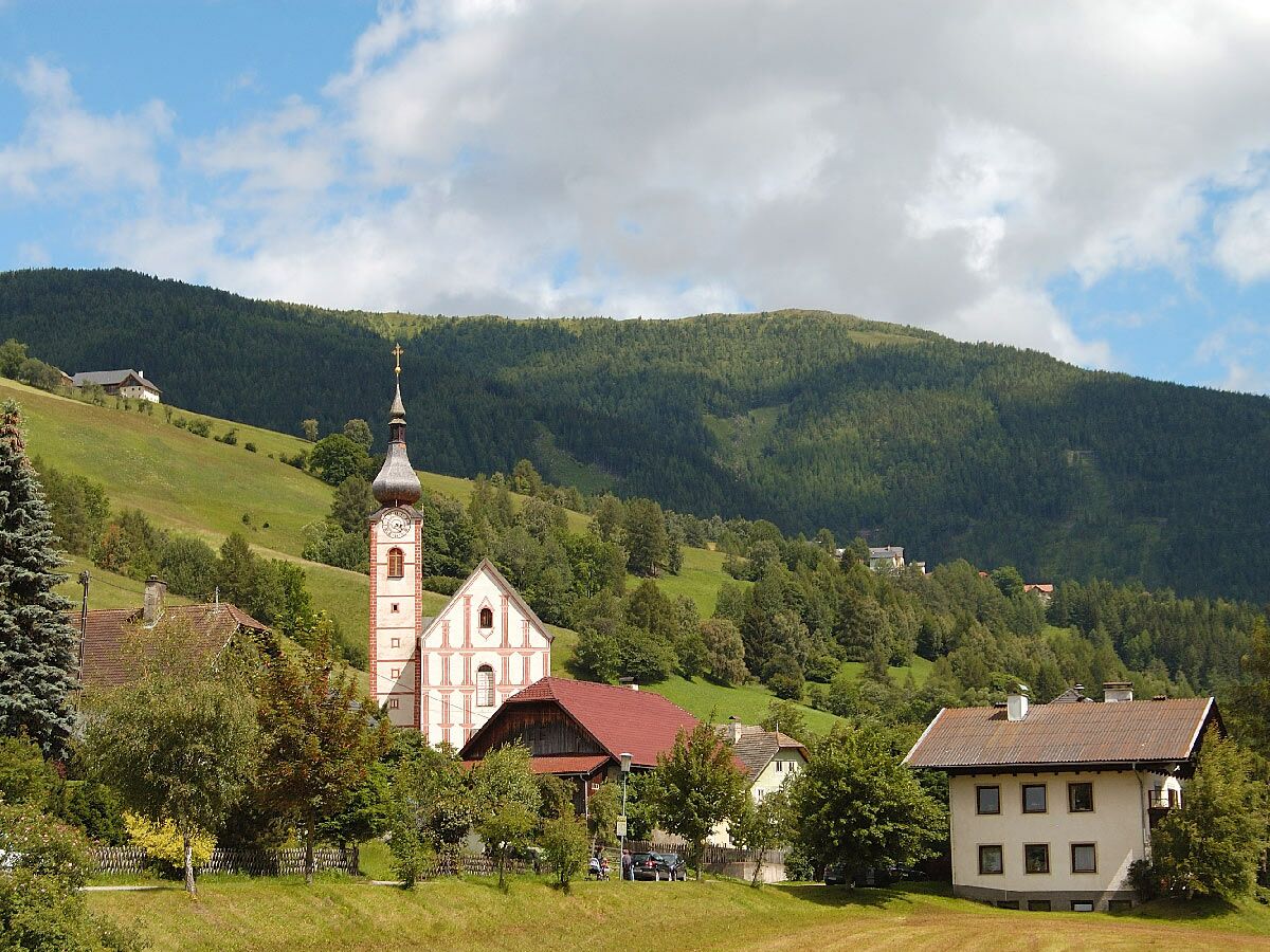 Malerisches Alpen Dorf mit einer rot-weißen Kirche und Zwiebelturm, umgeben von grünen Wiesen und bewaldeten Bergen.