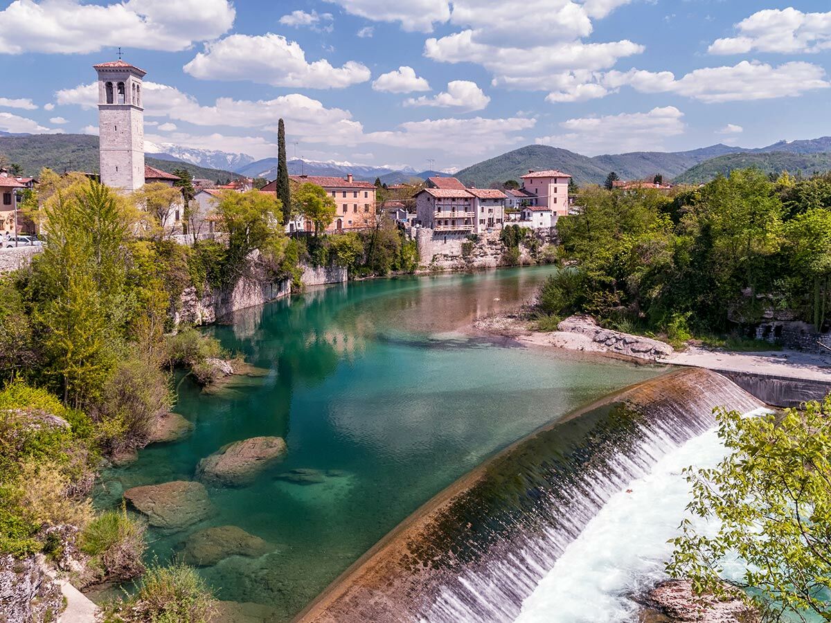 Malerische Stadt am türkisfarbenen Fluss mit Wasserfall, historischem Glockenturm und schneebedeckten Alpen im Hintergrund.
