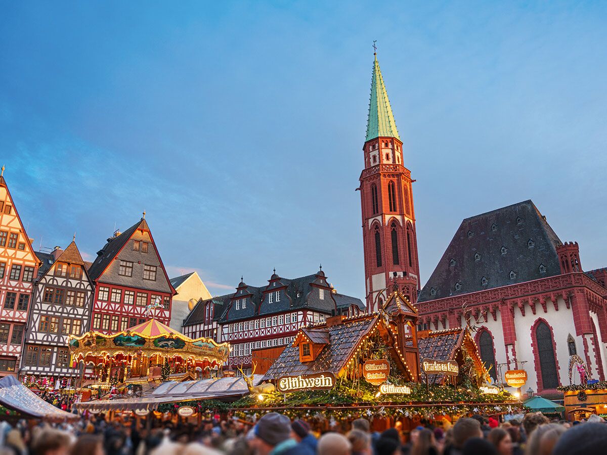 Beleuchteter Frankfurter Weihnachtsmarkt mit Glühweinständen, Karussell, Fachwerkhäusern und Paulskirche bei Dämmerung.
