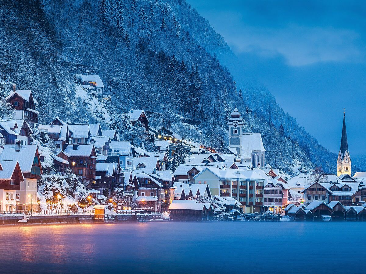 Malerisches Hallstatt Winterdorf am Seeufer mit verschneiten Häusern, Bergen und leuchtenden Lichtern in der Dämmerung.