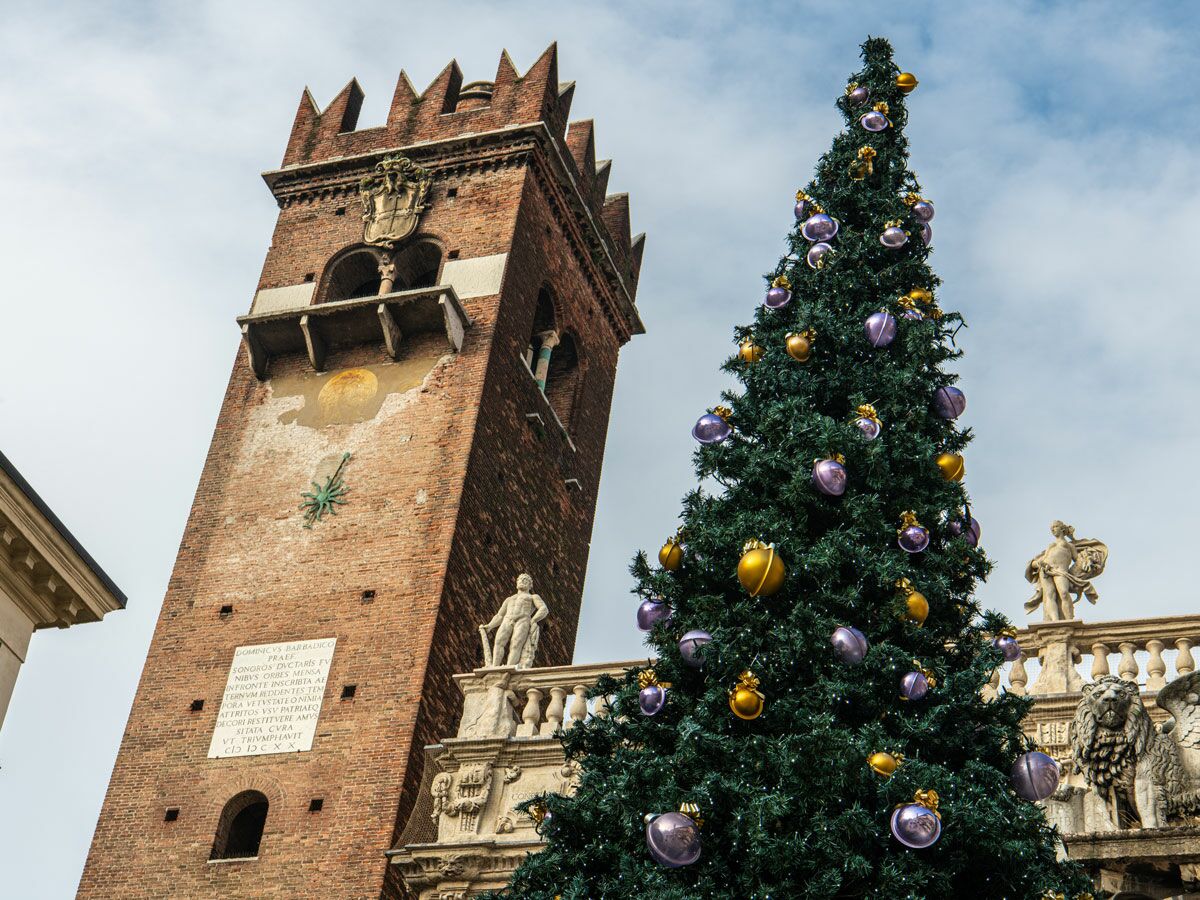 Großer Weihnachtsbaum mit goldenen und violetten Kugeln vor einem hohen, historischen Ziegelturm in Verona, Italien.