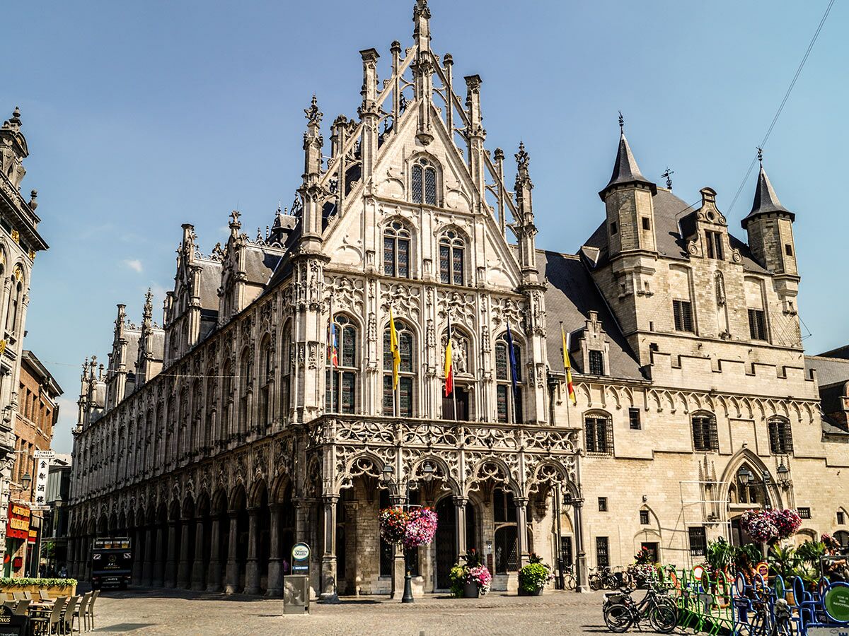 Die beeindruckende Tuchhalle von Ypern, ein gotisches Gebäude mit verzierten Fassaden und Fahnen, steht auf einem sonnigen Marktplatz.