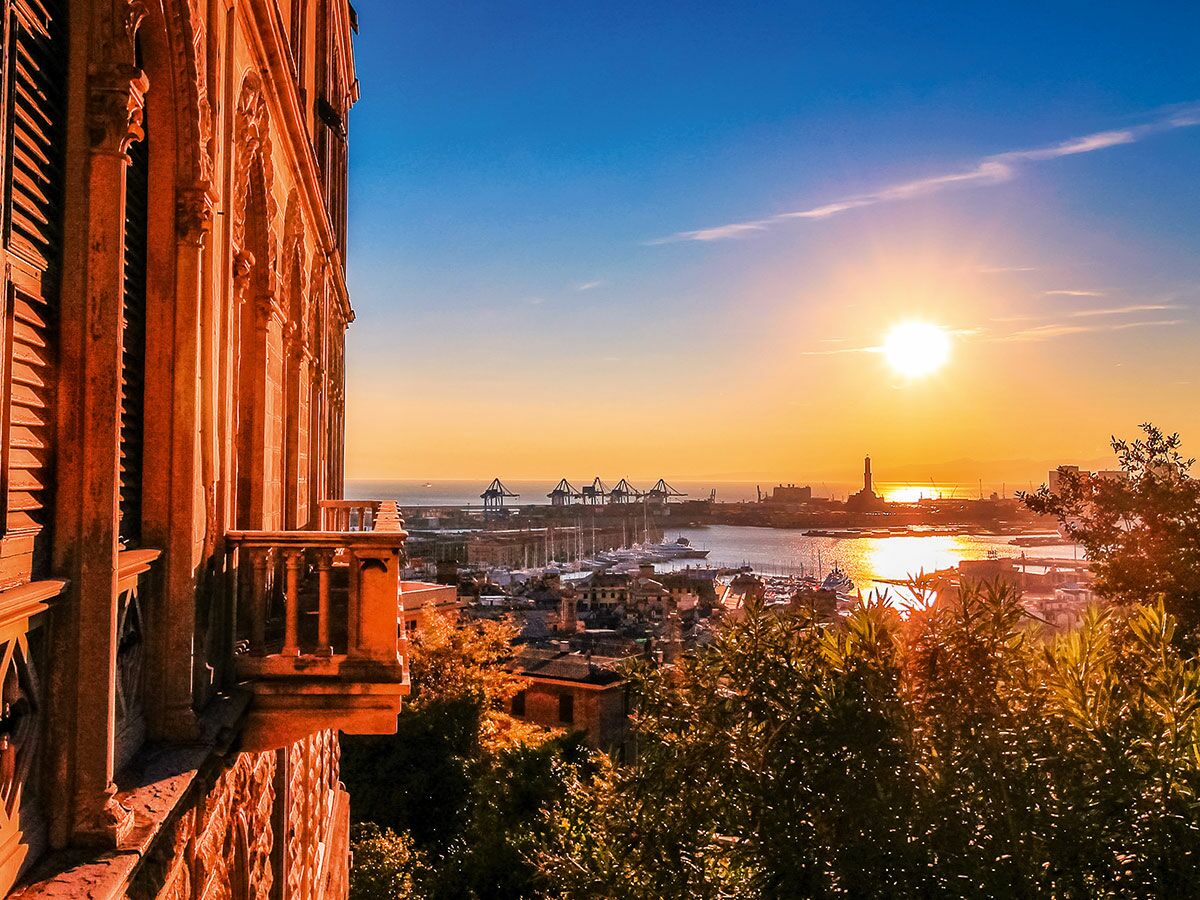 Goldener Sonnenuntergang über dem Hafen von Genua, gesehen von einem historischen Palast mit Balkon und Blick auf Leuchtturm und Boote.