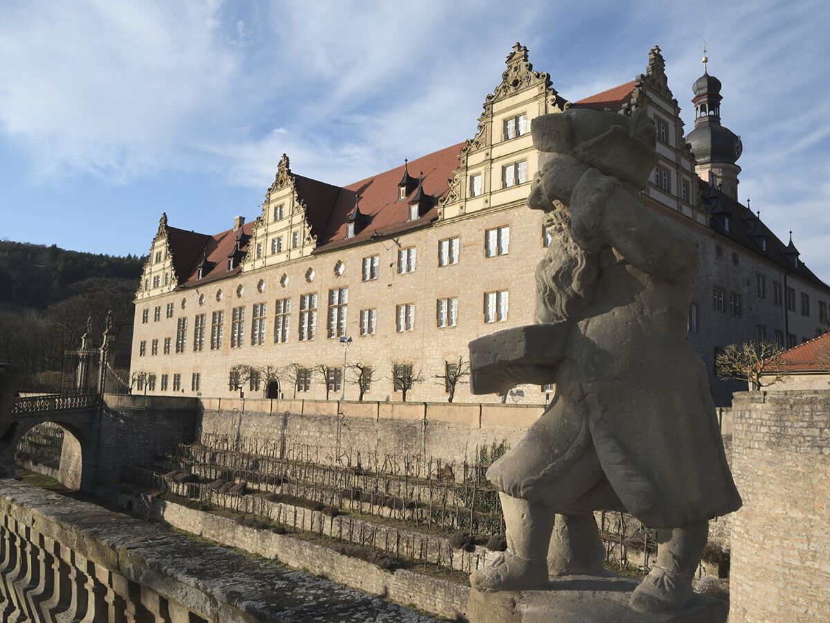 Historisches Schloss Weikersheim, Deutschland, mit detailreicher Barock-Architektur, vorgelagerter Stein-Statue, Brücke und Gartenanlage.