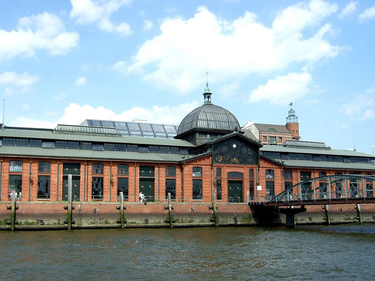 Die historische Fischauktionshalle in Hamburg mit Backsteinarchitektur, Kuppel und Brücke über dem Fluss unter blauem Himmel.