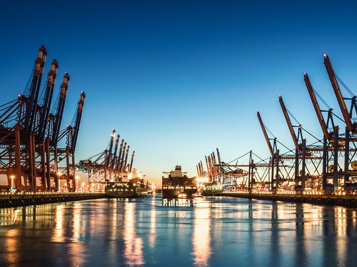Containerhafen Hamburg Burchardkai bei Nacht mit beleuchteten Kränen und Frachtschiffen, deren Lichter im Wasser spiegeln.