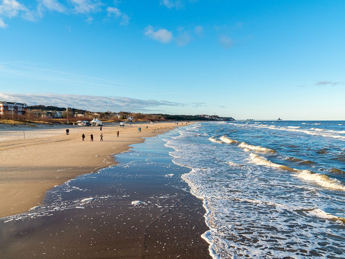 Weitläufiger Sandstrand mit spazierenden Menschen am Meer, sanfte Wellen, Küstenbebauung und blauer Himmel.