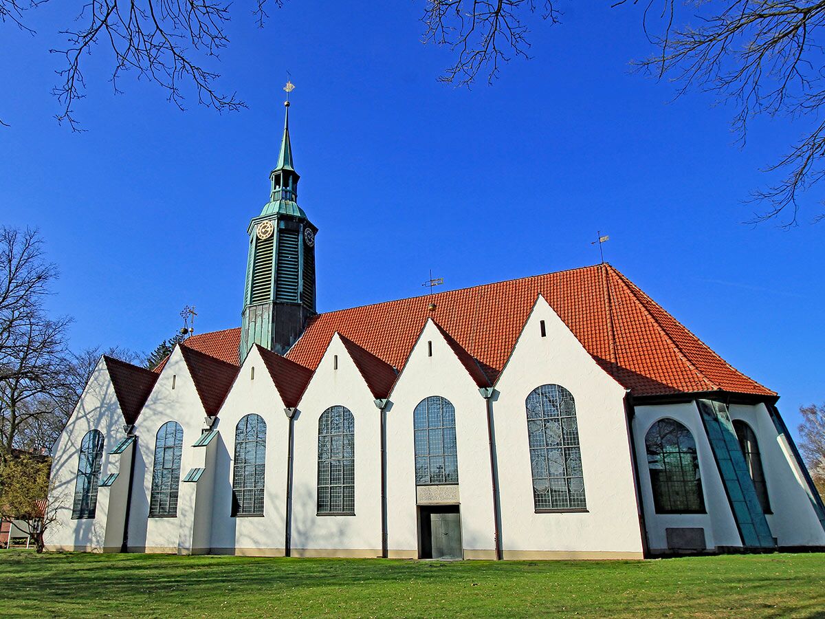 Sonnenbeschienene weiße Kirche mit rotem Ziegeldach, hohem Turm mit Uhr und Wetterfahne unter strahlend blauem Himmel.