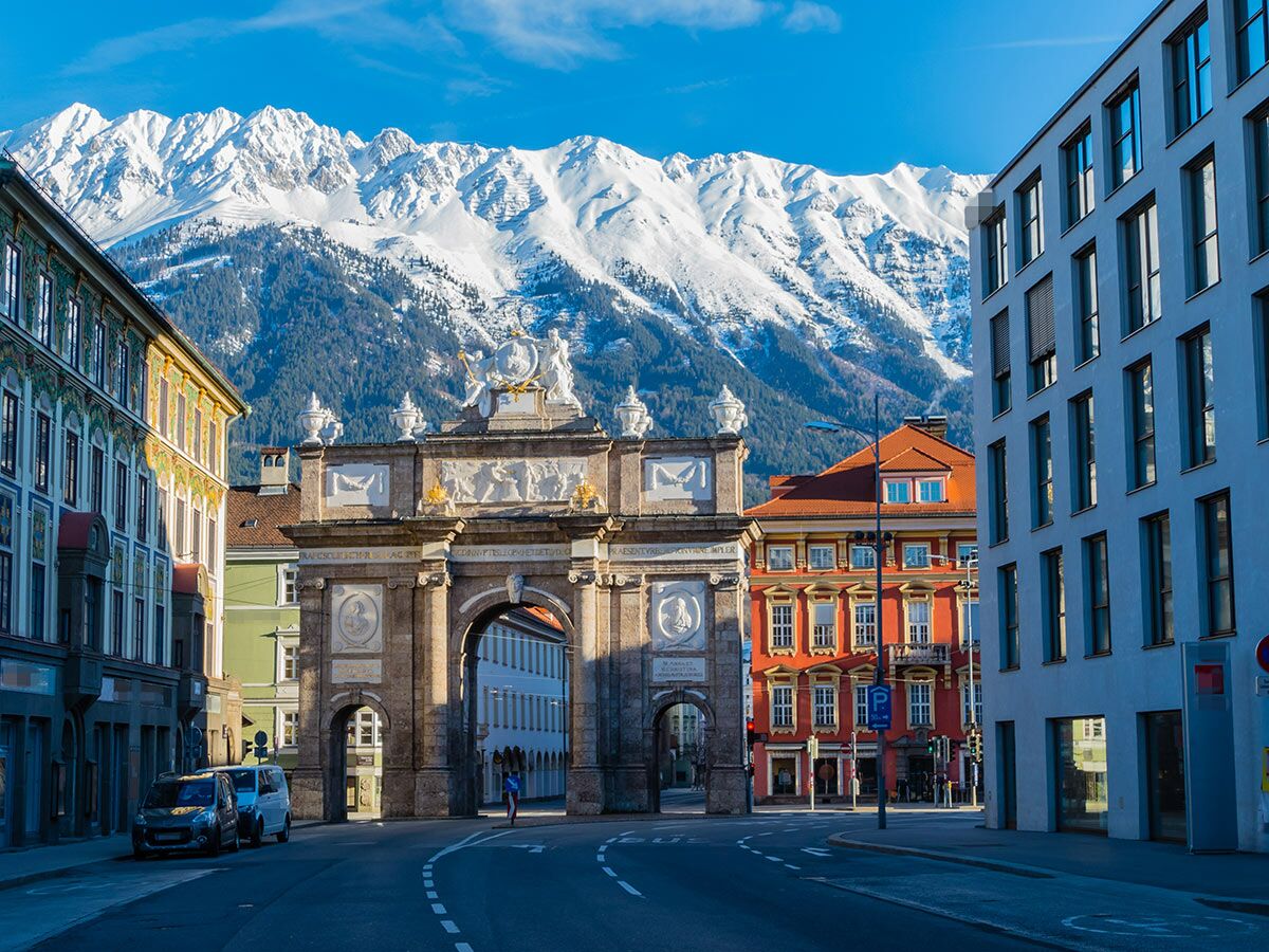 Triumphpforte Innsbruck: historische Architektur, moderne Gebäude, schneebedeckte Alpen und Straße unter blauem Himmel.