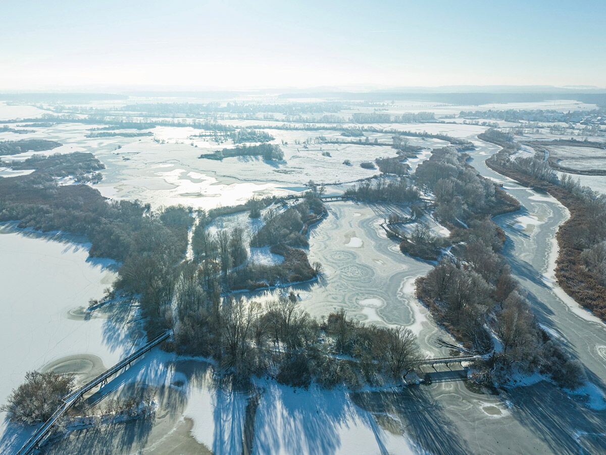 Luftaufnahme einer weiten, verschneiten Winterlandschaft mit einem gefrorenen Fluss, Inseln, Bäumen und Brücken.