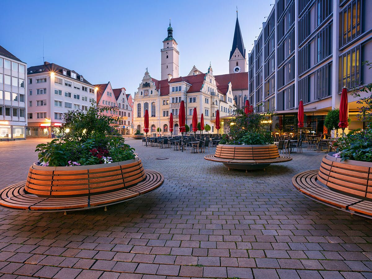 Panoramablick auf den historischen Altstadtplatz von Landshut in der Dämmerung mit modernen Sitzbänken, Cafés und beleuchteten Gebäuden.