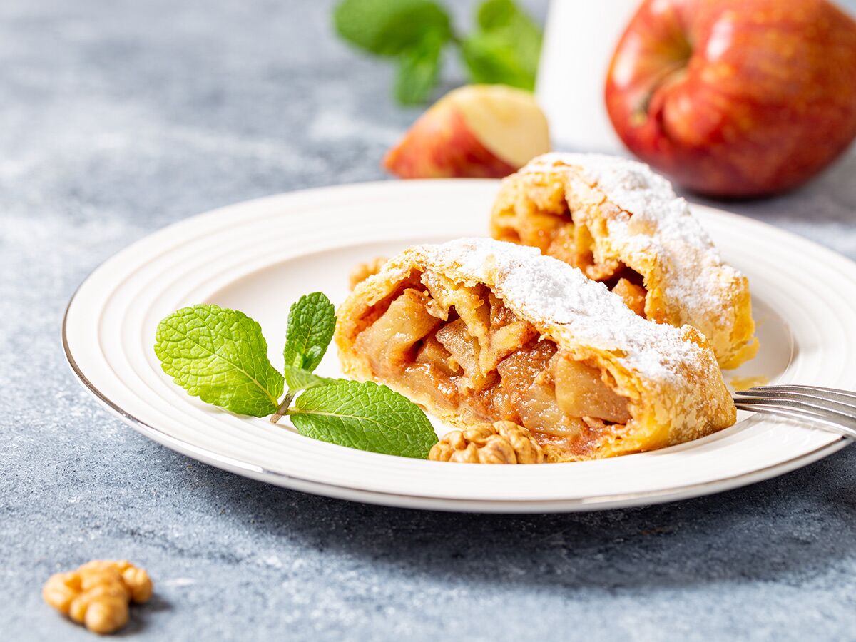Zwei Stücke Apfelstrudel mit Puderzucker, garniert mit Minze und Walnüssen auf einem weißen Teller, Äpfel im Hintergrund.