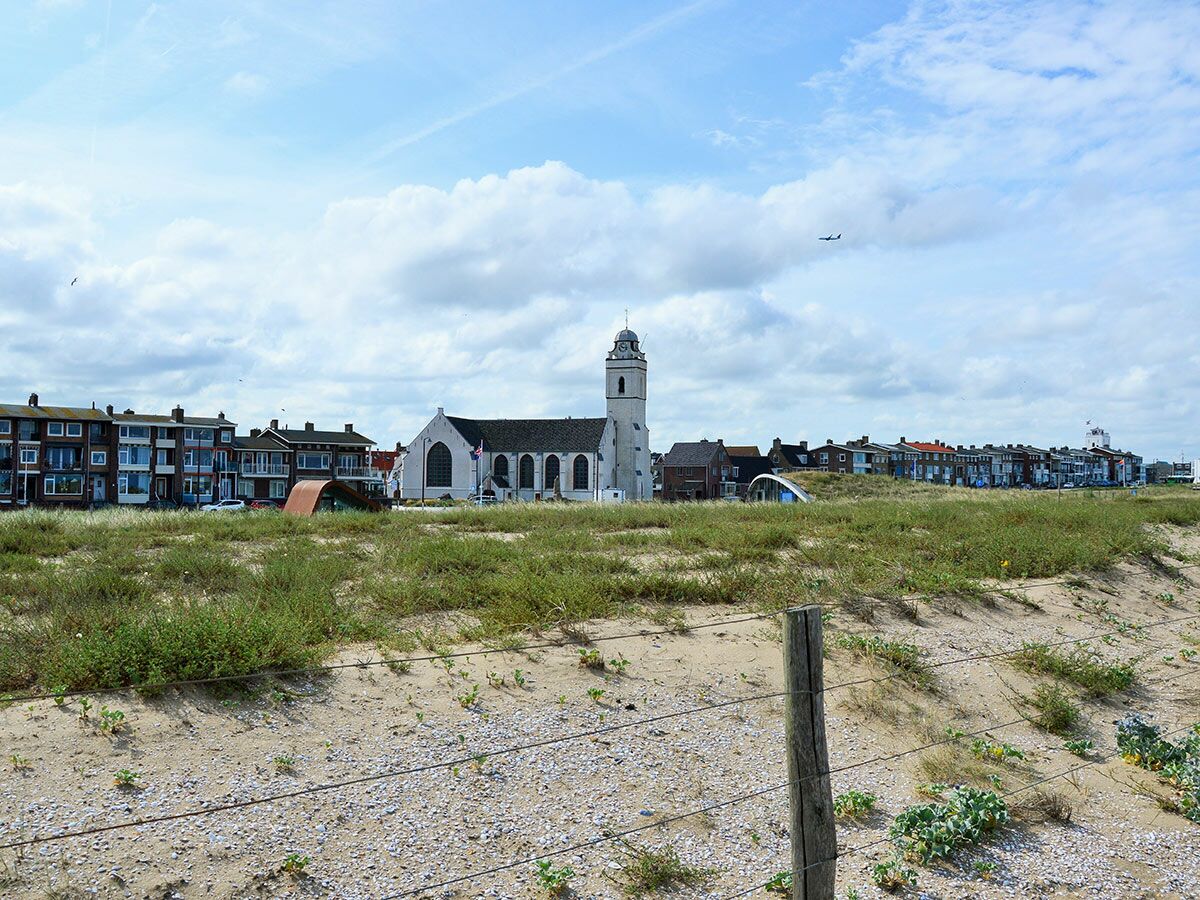 Blick über Sanddünen auf eine Küstenstadt mit markanter weißer Kirche, Häusern und blauem Himmel. Flugzeug fliegt vorbei.