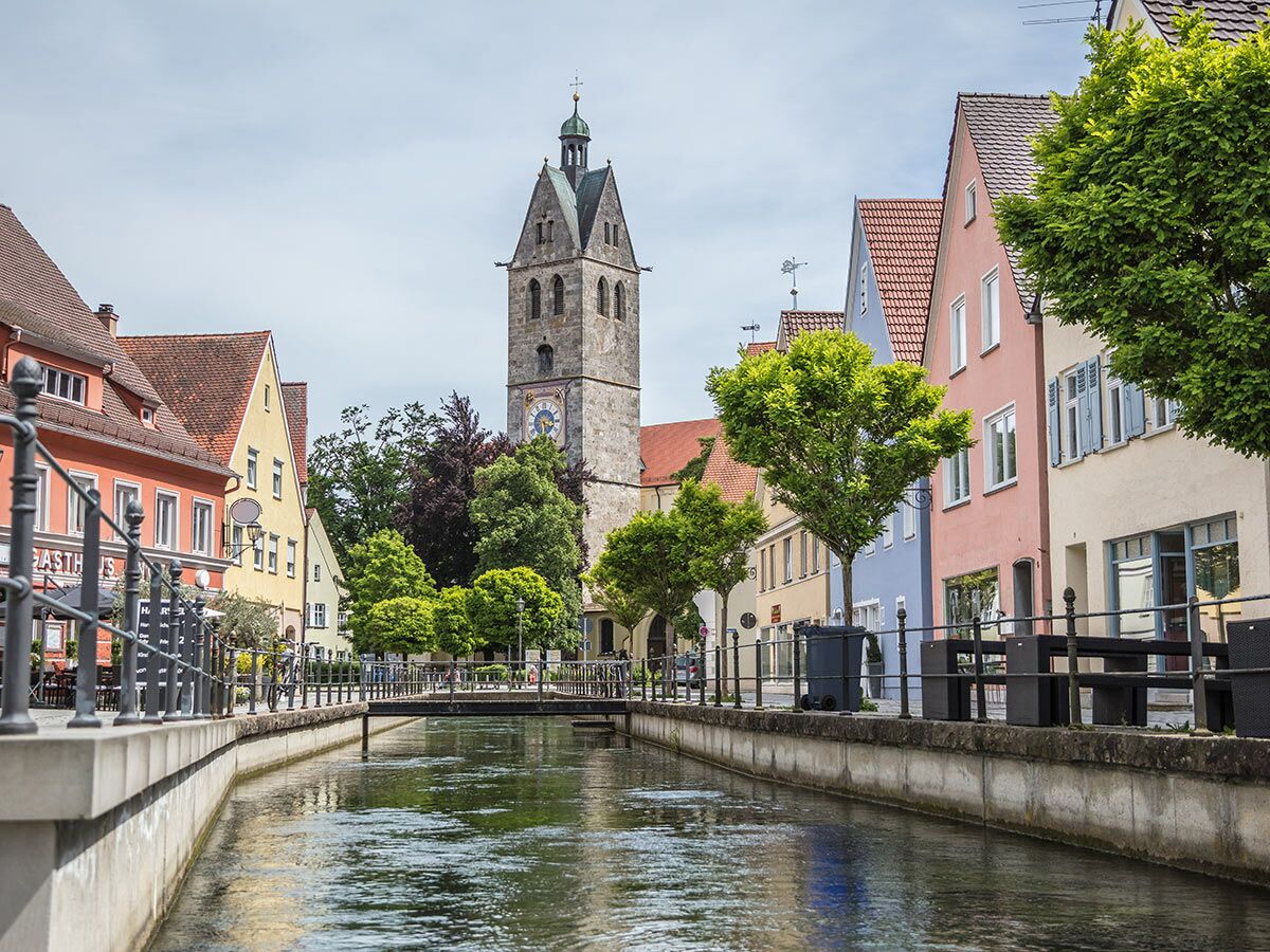 Blick auf eine historische deutsche Stadt mit bunten Häusern entlang eines Kanals, einem Kirchturm und grünen Bäumen.