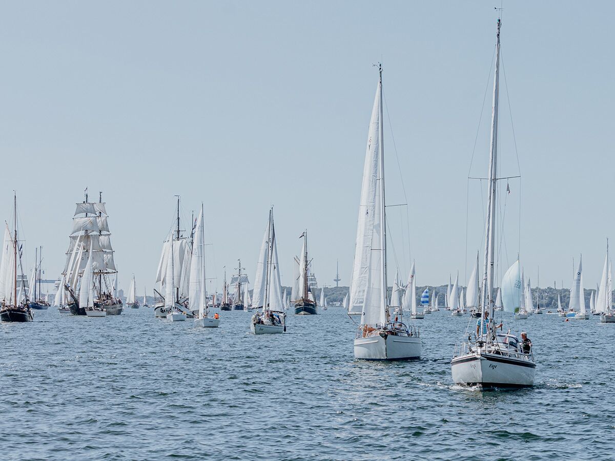 Zahlreiche Segelboote und historische Großsegler nehmen an einer sommerlichen Segelparade auf dem blauen Meer teil.