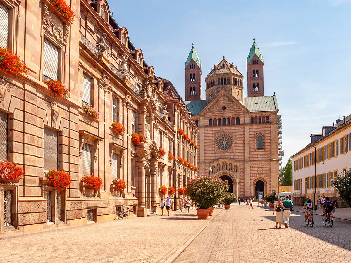 Der imposante Speyer Dom mit grünen Türmen an sonnigem Sommertag, umgeben von historischen Gebäuden und belebter Altstadtgasse mit Blumen und Menschen.