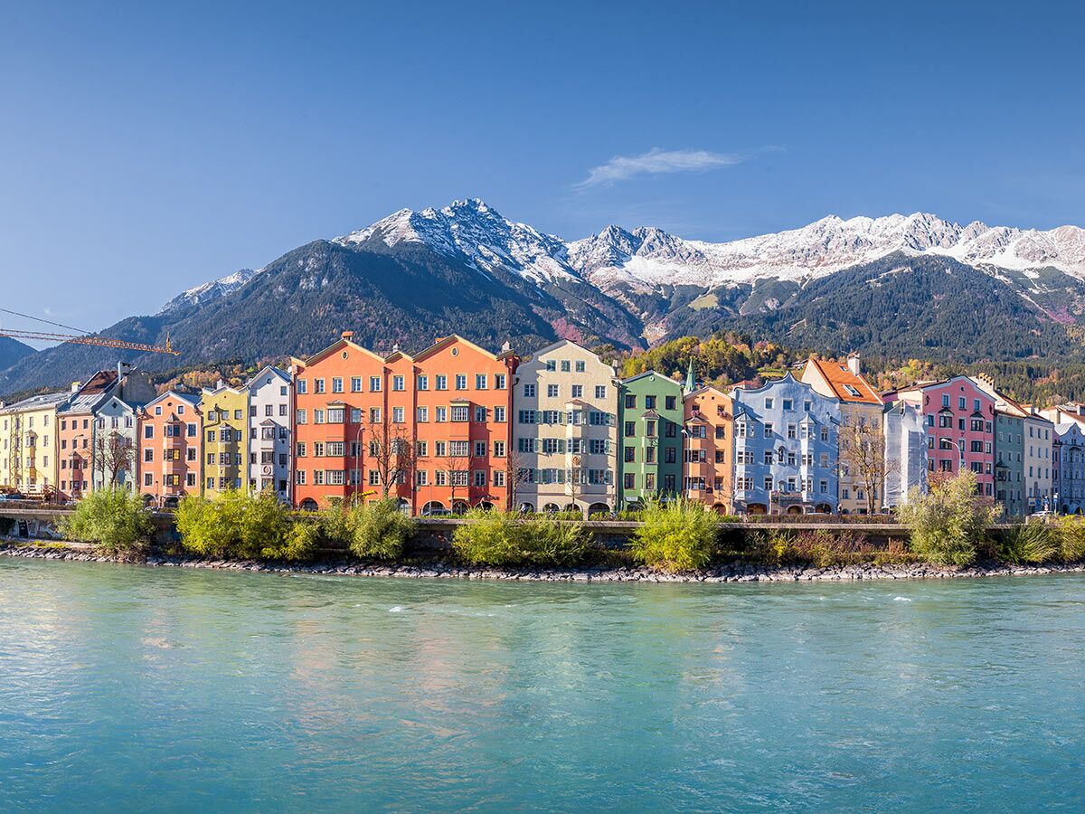 Blick auf bunte Häuser am Inn in Innsbruck mit schneebedeckten Alpen im Hintergrund unter blauem Himmel.